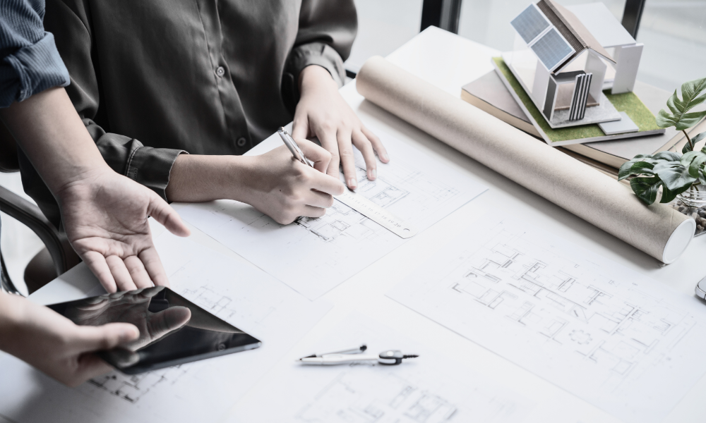 An engineering team is gathered around a table, intently reviewing technical drawings and project documents on various devices, including laptops and tablets. They are collaborating on sensitive project information and ensuring compliance with project deadlines while managing large files and maintaining version control in their architecture, engineering, and construction workflows.